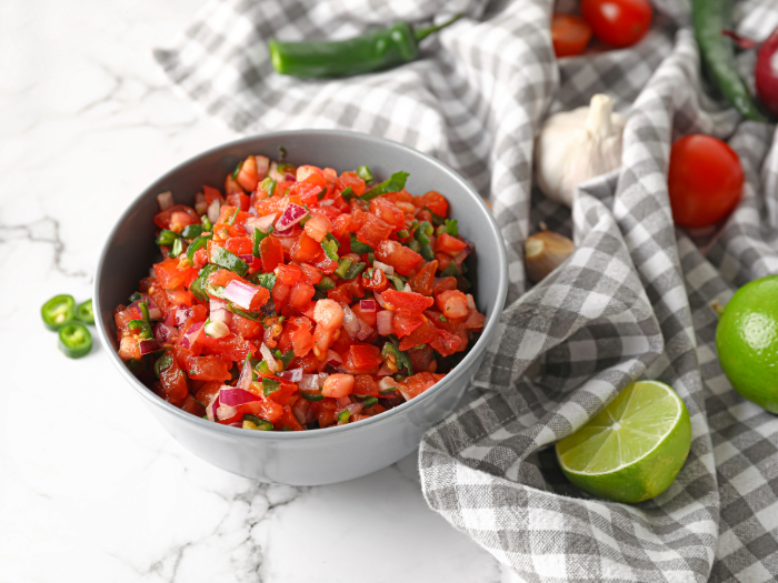 Tigela cinza com vinagrete, feito com tomate, cebola roxa, pimenta e coentro, cercado por limão, alho e pimentas sobre uma superfície de mármore com um pano xadrez.