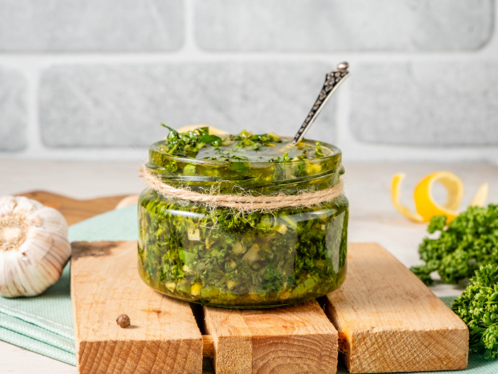 Pote de vidro com molho chimichurri caseiro, decorado com barbante rústico, apoiado sobre uma tábua de madeira, com alho e ervas frescas ao fundo.