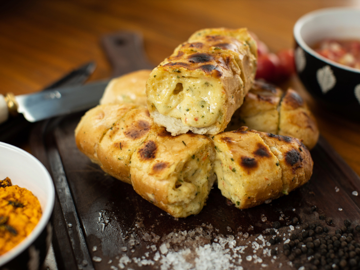 Pão de alho com queijo derretido e ervas, levemente tostado, servido sobre uma tábua de madeira com farofa e vinagrete.