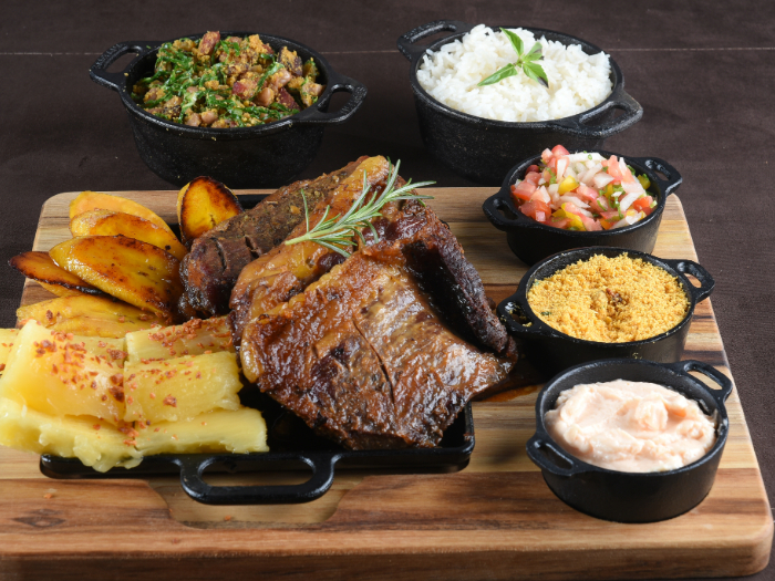 Tábua de churrasco assado, mandioca, batatas douradas e acompanhamentos como arroz, farofa, vinagrete, couve refogada e molho, servidos em pequenas panelas de ferro.