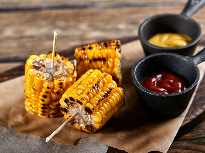 Espigas de milho grelhado cortadas em pedaços e espetadas com palitos, servidas sobre papel manteiga com molhos de ketchup e mostarda em mini panelas de ferro.