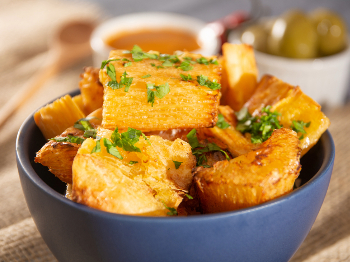 Pedaços de mandioca frita crocante, dourados e temperados com cheiro-verde, servidos em uma tigela azul, com molhos e azeitonas ao fundo.