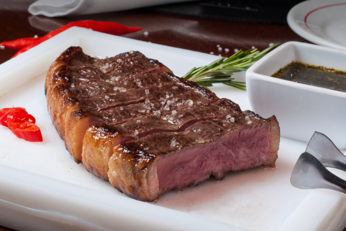 Um suculento bife de picanha assado ao ponto servido em uma tábua branca, com sal grosso por cima. Ao lado, ramos de alecrim, fatias de pimenta vermelha, um recipiente com molho e um garfo de churrasco. O fundo mostra um prato branco com detalhes vermelhos sobre uma mesa de madeira.