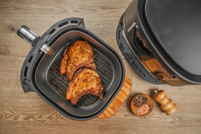 Dois pedaços de carne suína grelhados dentro de uma fritadeira elétrica, dispostos sobre uma grelha antiaderente. Ao lado, um moedor de pimenta, um recipiente com sal rosa e pimenta, e uma pequena tábua de madeira sobre uma superfície de madeira clara.