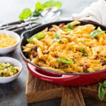 Macarrão gratinado com queijo e carne moída servido em uma frigideira vermelha de ferro fundido, decorado com folhas de manjericão. O prato está sobre uma tábua de madeira, acompanhado de potinhos com queijo ralado e picles, além de talheres e um guardanapo branco ao fundo.