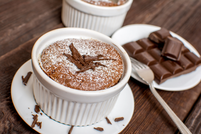 Suflê de chocolate servido em um ramequim branco, decorado com raspas de chocolate e açúcar de confeiteiro, acompanhado por um prato com pedaços de chocolate e um garfo sobre uma mesa de madeira rústica.