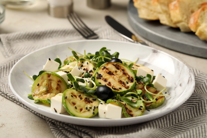 Salada com abobrinha grelhada, cubos de queijo branco, brotos, azeitonas pretas e sementes de gergelim preto servida em prato branco sobre guardanapo estampado.