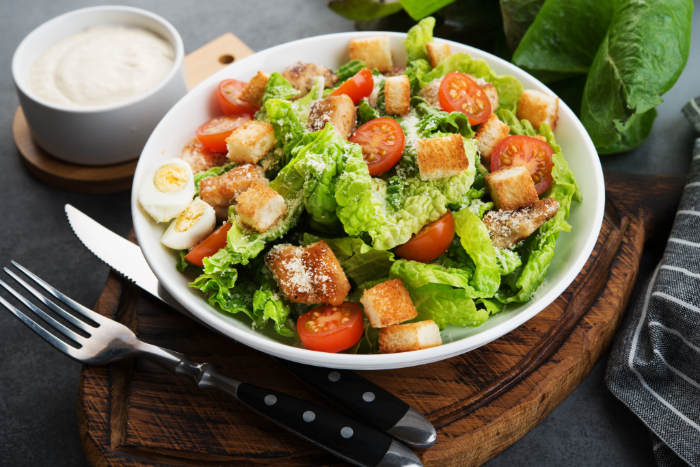 Salada Caesar servida em prato branco com alface romana, croutons, tomate-cereja, ovos cozidos e queijo ralado, acompanhada de molho em potinho e talheres sobre tábua de madeira.