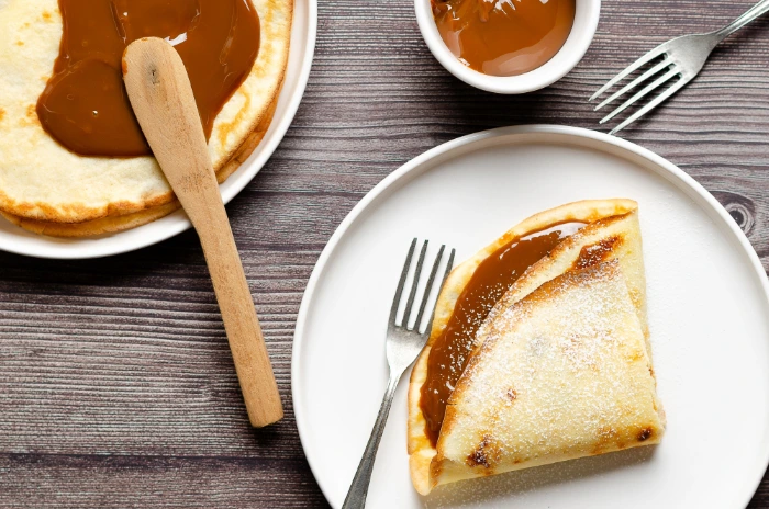 Panqueca dobrada recheada com doce de leite e polvilhada com açúcar, servida em prato branco sobre mesa de madeira, com talheres e recipiente de doce ao lado.