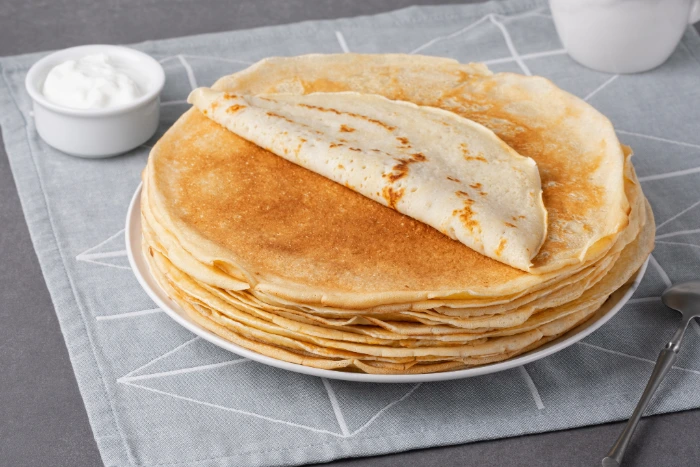 Pilha de panquecas douradas em prato branco, com uma panqueca parcialmente dobrada no topo, acompanhada por potinhos de creme ao fundo sobre uma toalha cinza.