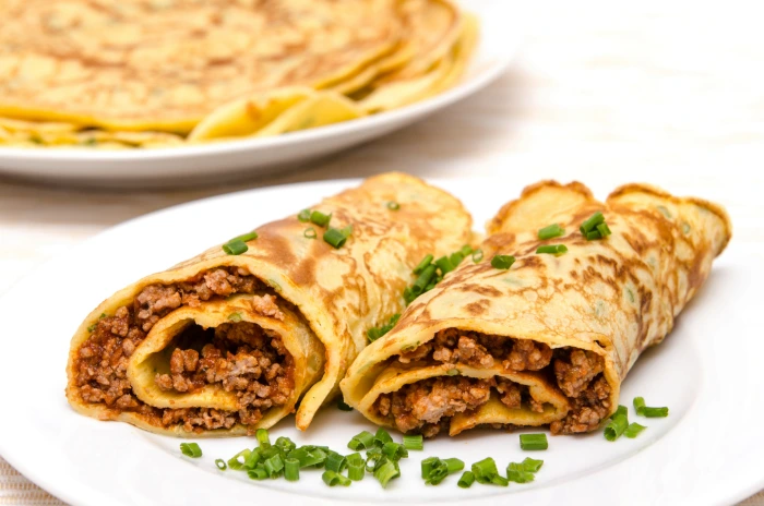 Panquecas recheadas com carne moída e cobertas com cebolinha, servidas em prato branco com mais panquecas ao fundo.