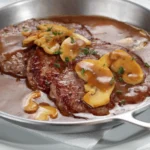 Prato de bife ao molho madeira servido em panela de inox, com fatias de cogumelo e molho espesso, acompanhado de salsinha picada por cima.