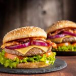 Dois hambúrgueres artesanais com queijo, bacon, alface, tomate e cebola roxa, servidos em pão com gergelim sobre pratos de pedra, com fundo rústico de madeira.