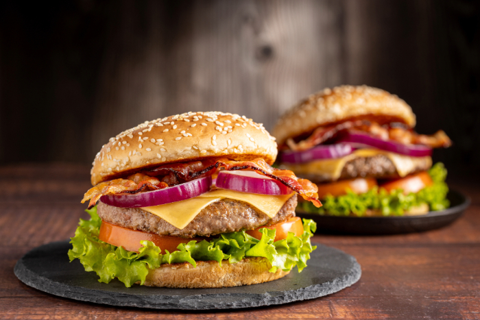 Dois hambúrguers artesanais com pão de gergelim, hambúrguer bovino, queijo derretido, bacon crocante, cebola roxa, alface e tomate servidos em pratos de ardósia sobre mesa de madeira rústica