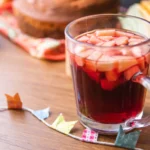 Caneca de quentão de vinho com pedaços de maçã, decorada em mesa de madeira com bolo, milho e bandeirinhas de festa junina ao fundo.