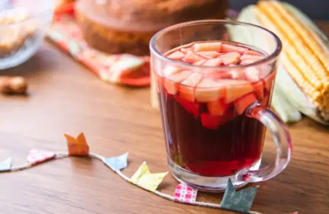 "Xícara de vidro transparente com quentão de cor avermelhada servido quente, acompanhada de saquinhos de especiarias coloridos sobre mesa de madeira, com pães ao fundo