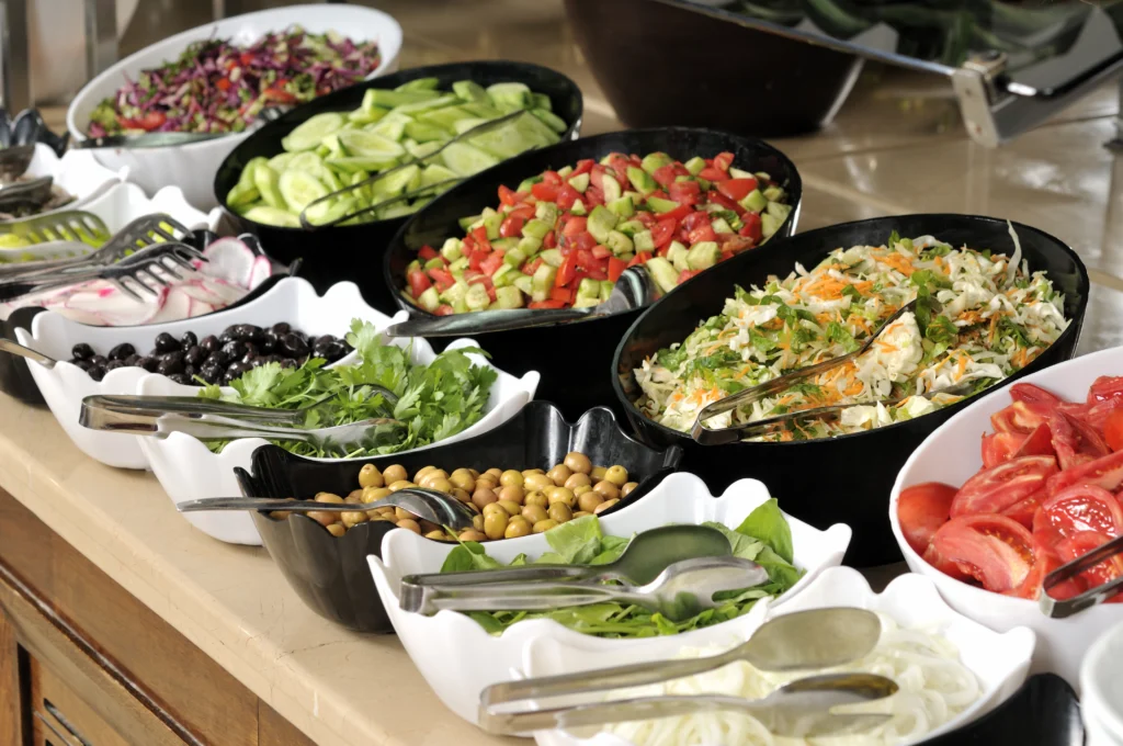 Buffet de saladas com tigelas de azeitonas, folhas verdes, pepino, tomate, grão-de-bico, repolho e outros legumes frescos