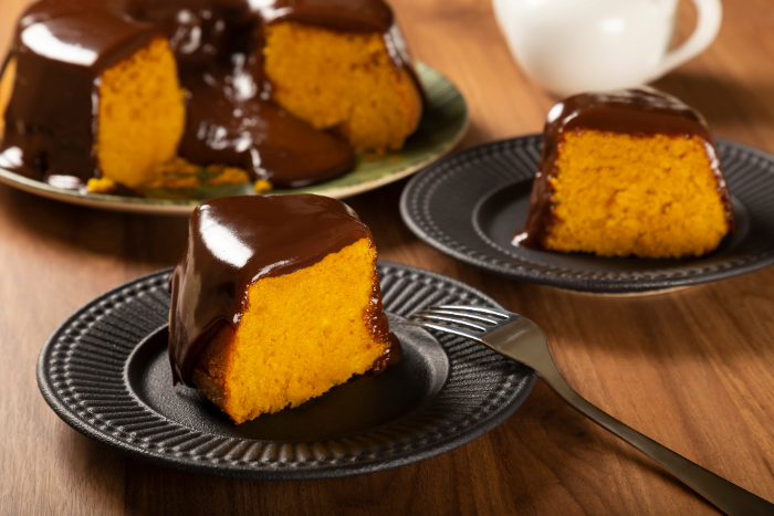Fatia de bolo de cenoura com cobertura de chocolate cremosa servida em prato preto sobre mesa de madeira, com o bolo inteiro ao fundo.
