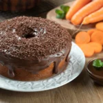 Bolo de cenoura com cobertura de chocolate e granulado servido em prato branco, ao lado de cenouras frescas fatiadas e uma tigela com calda de chocolate.