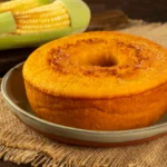 Bolo de milho cremoso inteiro com furo central em prato de cerâmica, ao lado de espigas de milho verde