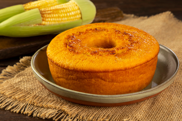 Bolo de milho cremoso inteiro com furo central em prato de cerâmica, ao lado de espigas de milho verde