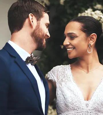 Noivo de terno azul marinho com gravata borboleta e noiva de vestido branco rendado com brincos de pérola se olhando carinhosamente e sorrindo durante cerimônia de casamento ao ar livre com vegetação desfocada ao fundo