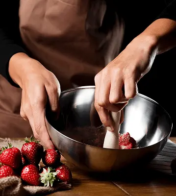 Mãos de chef derretendo chocolate escuro em tigela de inox sobre banho-maria, com morangos frescos vermelhos dispostos sobre tábua de madeira ao lado, preparando sobremesa gourmet na cozinha