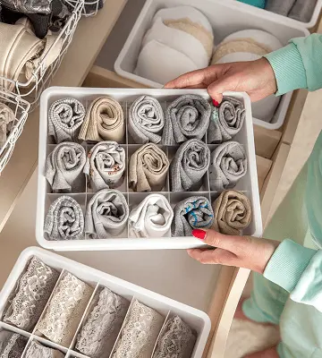 Mãos femininas organizando gaveta com divisórias contendo roupas íntimas e meias enroladas de forma organizada, com caixas organizadoras cinzas e outros compartimentos para acessórios, demonstrando método de organização doméstica