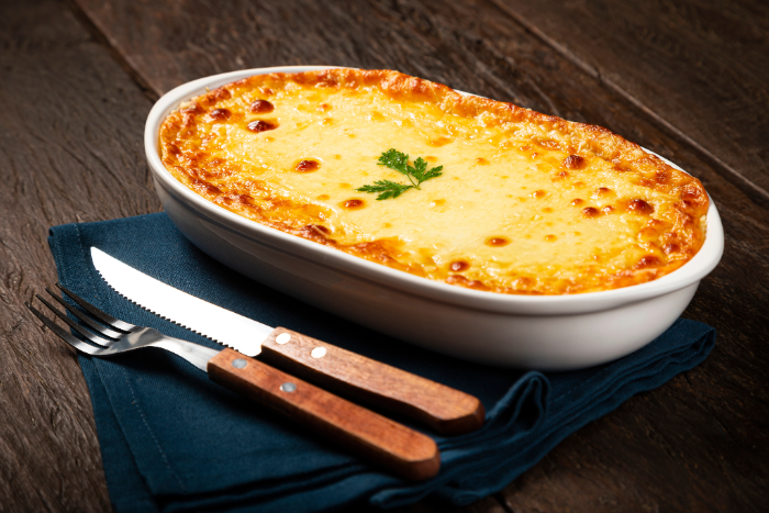 Escondidinho de carne moída com purê de batata e queijo gratinado em travessa branca, decorado com salsinha, sobre guardanapo azul e talheres de madeira.