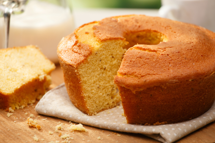Bolo de fubá com erva-doce simples, em formato redondo com furo central, sobre pano de algodão estampado, com fatia cortada ao lado e farelos espalhados sobre uma tábua de madeira. Ao fundo, jarra de leite desfocada.