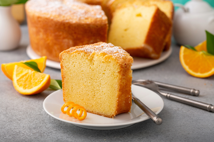 Fatia de bolo de laranja fofinho em prato branco com raspas de casca ao lado, acompanhada de garfo, com o bolo inteiro ao fundo, decorado com açúcar de confeiteiro e fatias de laranja frescas ao redor.