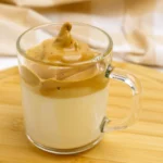 Copo de vidro com leite gelado e creme de café batido por cima, formando o café cremoso pronto para servir.