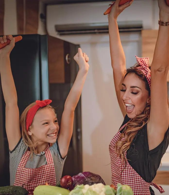 Mãe e filha com aventais vermelhos e fitas no cabelo, sorrindo e levantando os braços em celebração enquanto preparam vegetais frescos na cozinha
