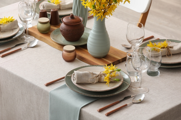 Mesa posta elegante com pratos em tons de verde, vasos decorativos, flores amarelas, taças de vidro, guardanapos de tecido e talheres sobre toalha clara.